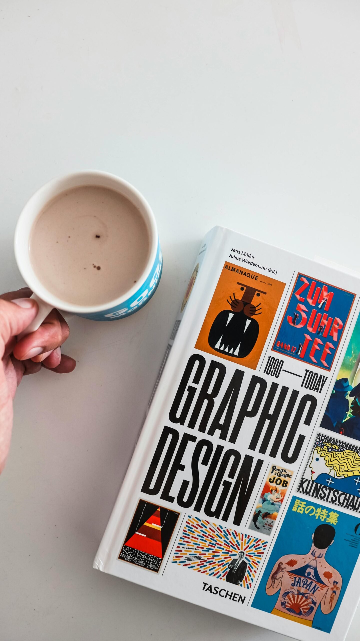 Close-up of a hand holding a coffee cup next to a graphic design book on a white surface.